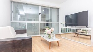 A modern living room with a white sofa, a coffee table with a vase of flowers, a large flat-screen TV, and a glass partition.