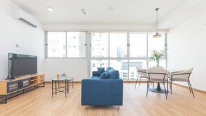Un salon moderne avec un canapé bleu, un téléviseur, une table à manger avec des chaises et de grandes fenêtres donnant sur la ville.