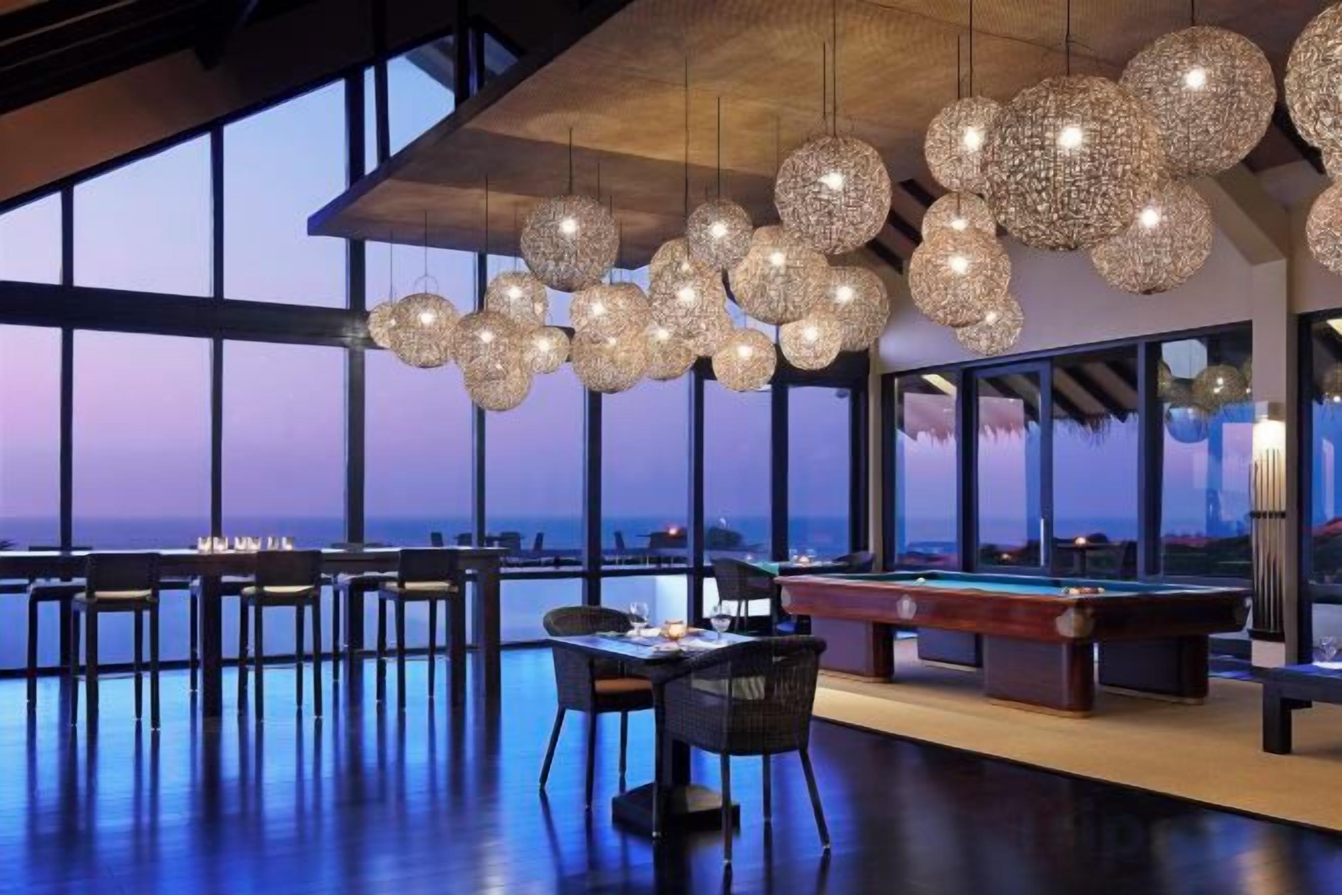 Un vaste espace intérieur comprenant une table de billard, des tables et des chaises, de grandes fenêtres et des luminaires sphériques suspendus.
