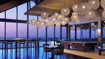 A spacious indoor area with a pool table, dining tables, and chairs, featuring large windows and hanging spherical light fixtures.