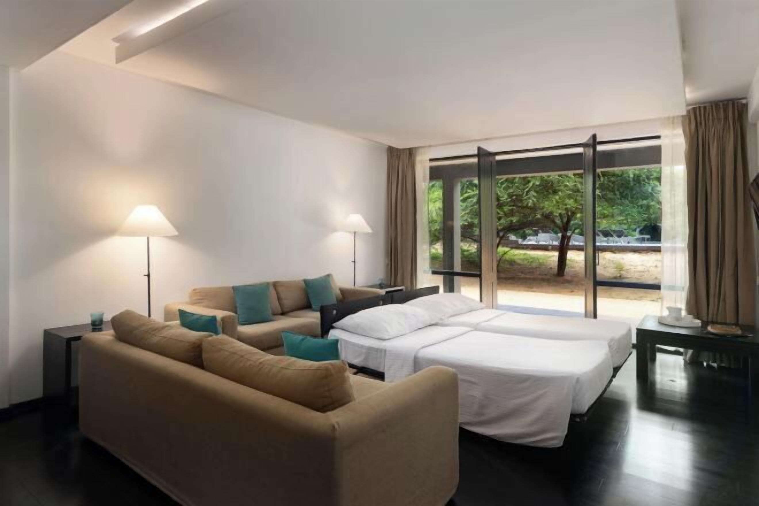 A hotel room with a sofa, two beds, a lamp, and a view of greenery outside.