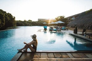 A poolside area with a person sitting by the pool, lounge chairs, umbrellas, and a thatched-roof structure. - Jetwing Yala (Palatupana)