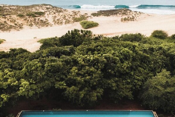 Una piscina rodeada de abundante vegetación y una playa arenosa con olas del océano al fondo.