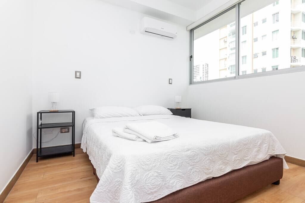A hotel room with a bed, a nightstand, an air conditioning unit, and a window with a view of buildings.