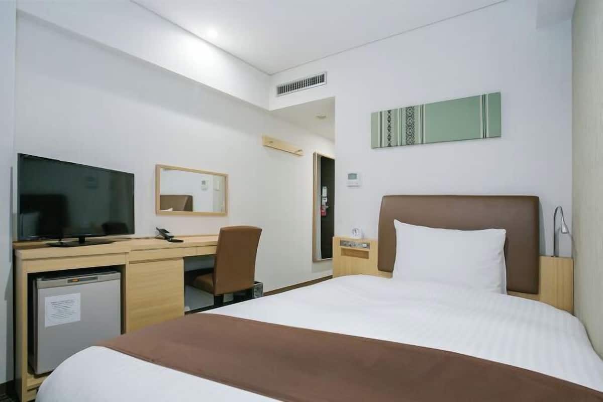 Una habitación de hotel con cama, escritorio, televisor, espejo y un refrigerador pequeño.