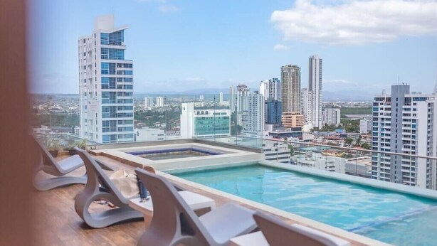 Una piscina en la azotea con sillones de descanso, vista a un paisaje urbano con rascacielos y cielo despejado.