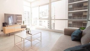 Una sala de estar moderna con un sofá, una mesa de centro y un televisor sobre un mueble de madera. Amplias ventanas ofrecen vistas a la ciudad.