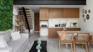 A modern living space with a sofa, coffee table, and wicker chairs. A kitchen area is visible with wooden cabinets and a white countertop. - Moderno Loft Temple II A112 By Sandy Powerful (Tulum)