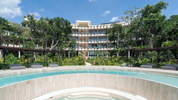 A swimming pool area with a curved design, surrounded by lush greenery and a multi-story building in the background. - Moderno Loft Temple II A112 By Sandy Powerful (Tulum)