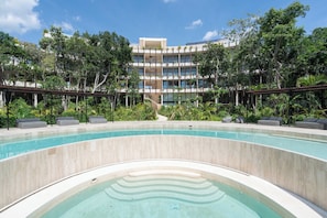 Un área de piscina con diseño curvo, rodeada de exuberante vegetación y un edificio de varios pisos al fondo.