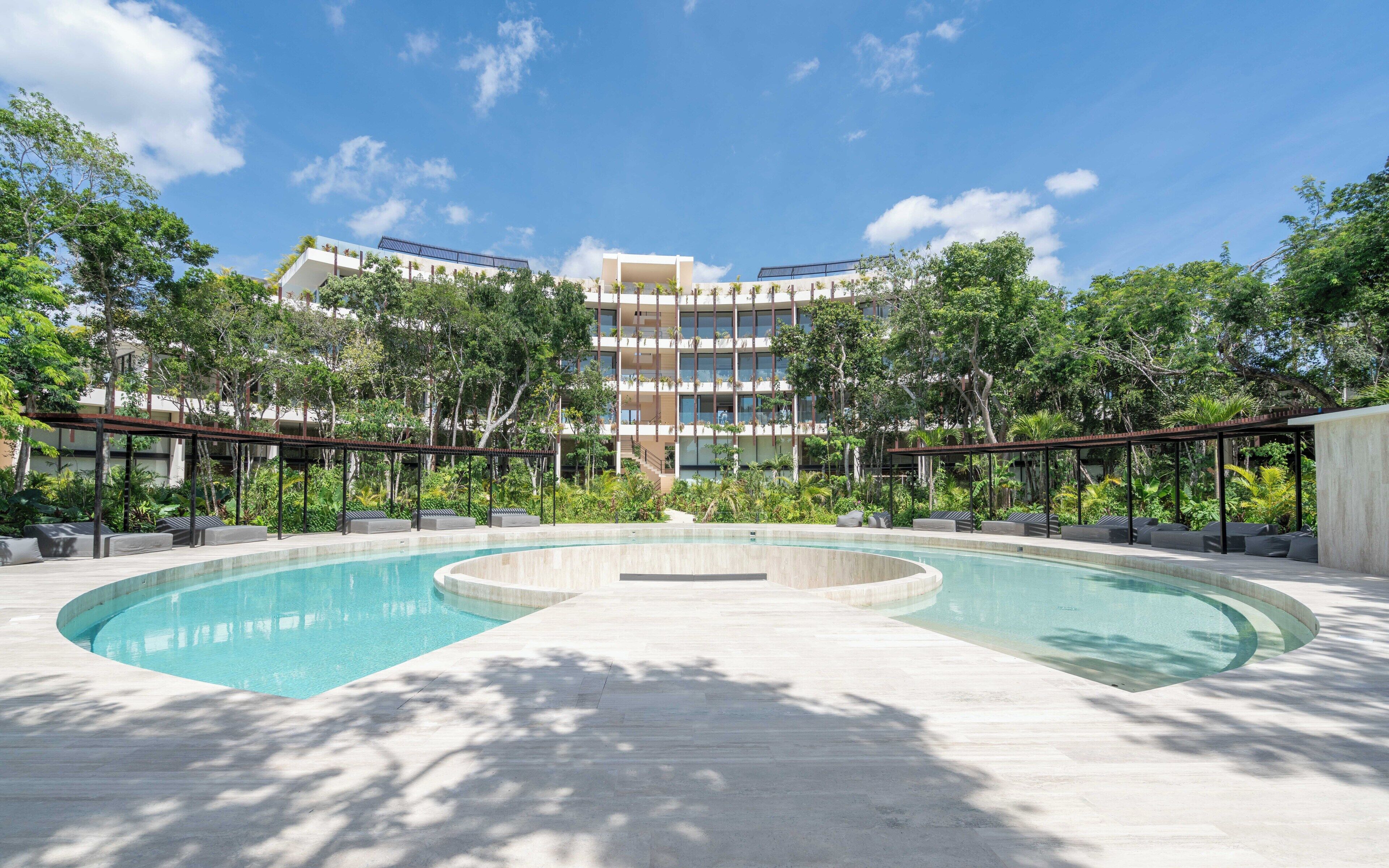 A modern hotel with a large pool, surrounded by lush greenery and a multi-story building with balconies.