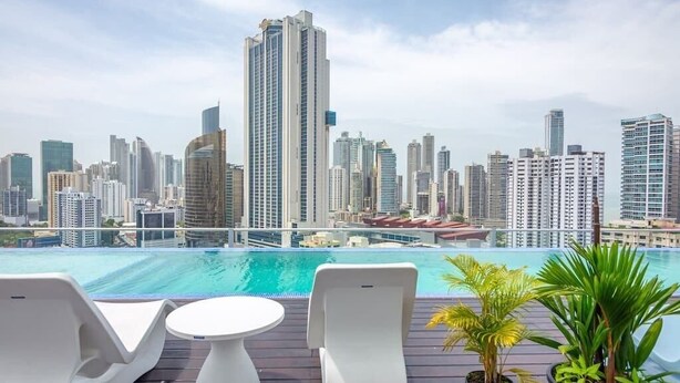A rooftop pool area with lounge chairs and a table, overlooking a city skyline with high-rise buildings.