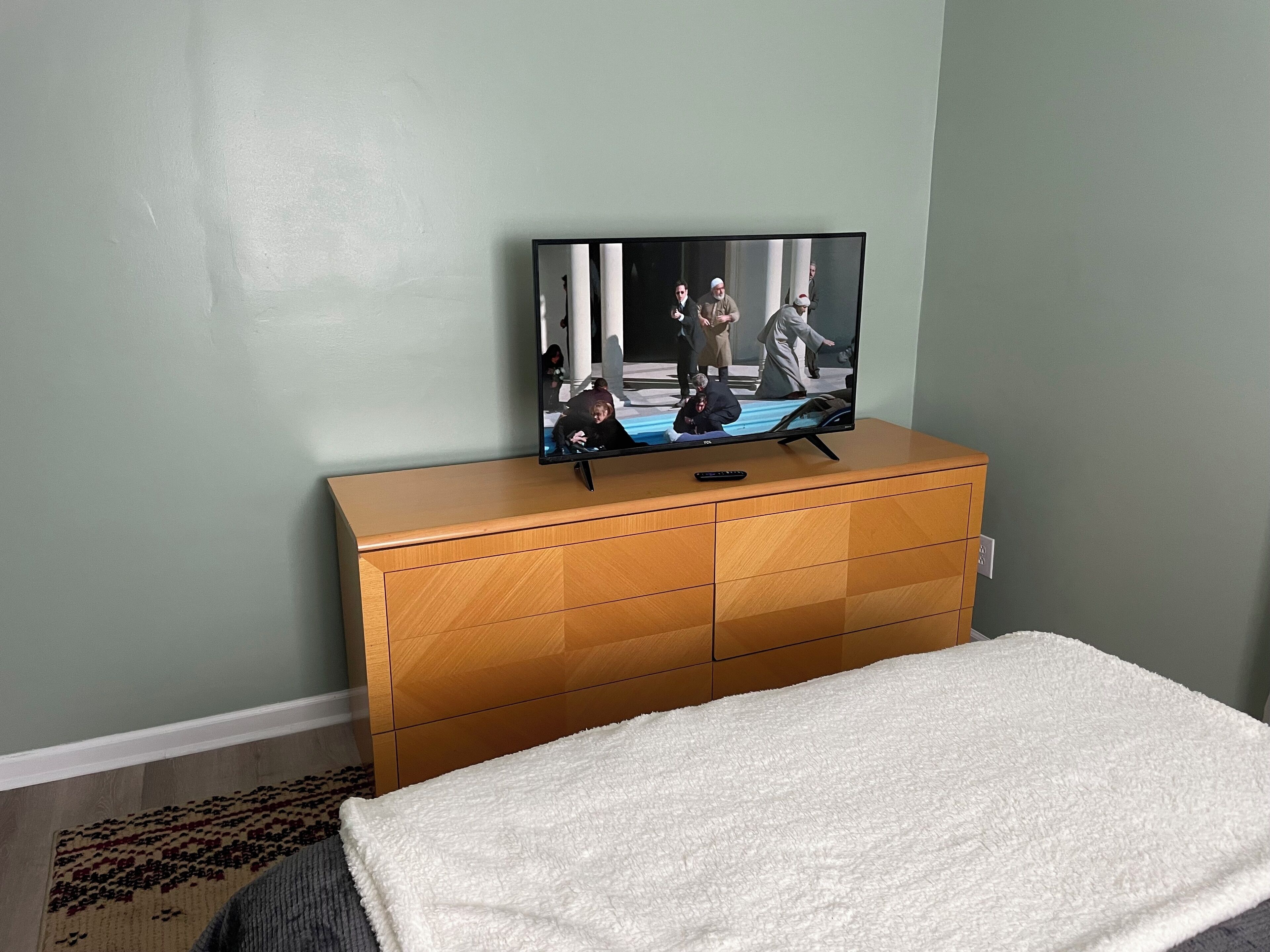 A TV on a wooden dresser in a room with a white carpet and light green walls.
