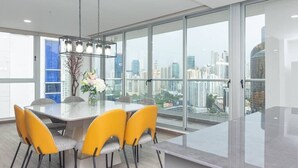 A modern dining area with a large table and yellow chairs, large windows with a city view, and a hanging light fixture.