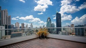 A rooftop terrace with a wooden floor and potted plants, overlooking a cityscape with modern buildings and a clear sky.