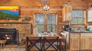 A rustic cabin interior with a stone fireplace, wooden dining table, and a kitchen area. - You need a Time Out on Your Next Getaway? (Sevierville)