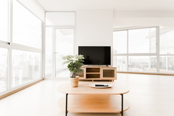 Ein modernes Wohnzimmer mit großem Fenster, Flachbildfernseher, Couchtisch aus Holz und einer Topfpflanze.
