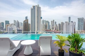 A rooftop pool area with lounge chairs and a table, overlooking a city skyline with high-rise buildings.