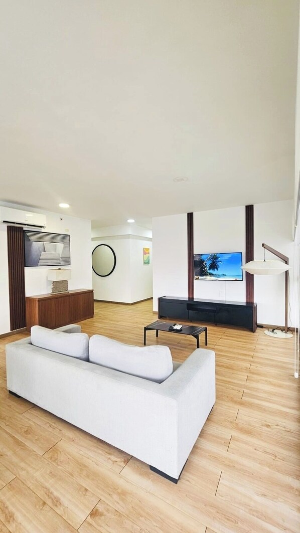A modern living room with a white sofa, a flat-screen TV mounted on the wall, a wooden floor, and a small side table.