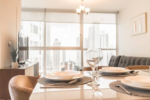 Une table à manger dressée avec des assiettes, des verres et des couverts, dans une pièce moderne dotée d’une grande fenêtre offrant une vue sur la ville.