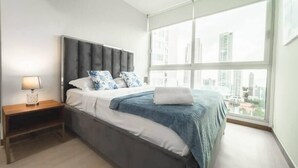 A modern bedroom with a large bed, a nightstand, a lamp, and a city view through large windows.