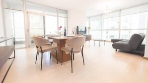 A spacious living area with a dining table, chairs, a sofa, and large windows providing natural light.