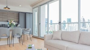 A modern living room with a sofa and dining area, large windows with a city view, and a kitchen in the background.