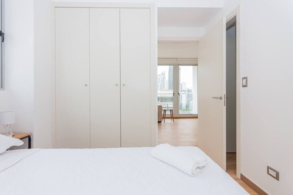A hotel room with a large wardrobe, a bed with a rolled-up white towel, a nightstand with a lamp, and a view of the city through large windows.