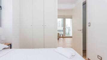 A hotel room with a large wardrobe, a bed with a rolled-up white towel, a nightstand with a lamp, and a view of the city through large windows.