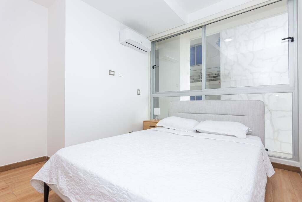 A modern bedroom with a large bed, a nightstand, a wall-mounted air conditioner, and a large window with a stone-like pattern.