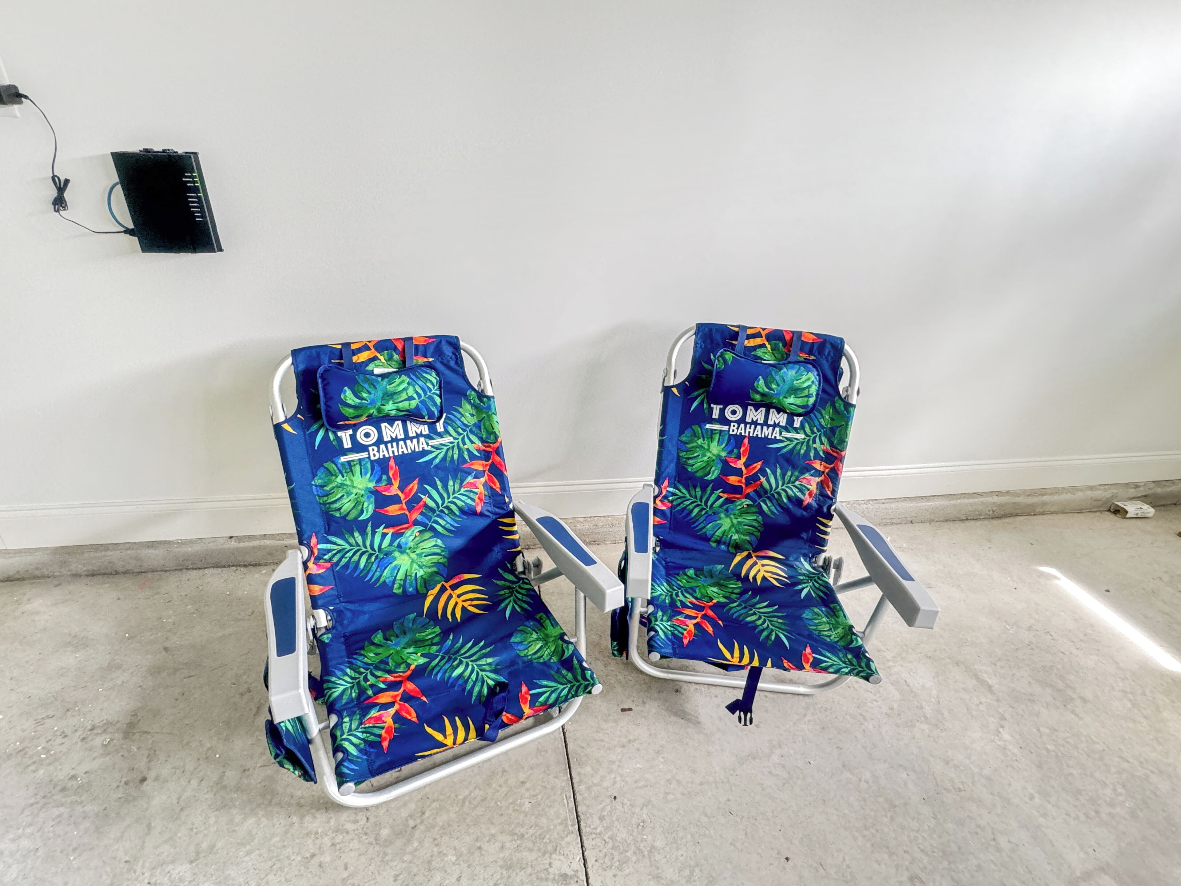 Two reclining chairs with a tropical leaf pattern and 'TOMMY BAHAMA' branding, positioned against a plain wall in a room with a concrete floor.