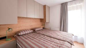 A bedroom with a wooden headboard, a bed with striped bedding, a nightstand with a lamp, and a window with curtains. - Apartment Martinus-r (Sveti Martin na Muri)