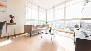 Ein modernes Wohnzimmer mit einer Couch, einem Couchtisch und einem Schreibtisch. Große Fenster bieten Blick auf die Stadt.