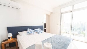 Ein Hotelzimmer mit Bett, Nachttisch, Lampe und Blick auf die Stadt durch große Fenster.