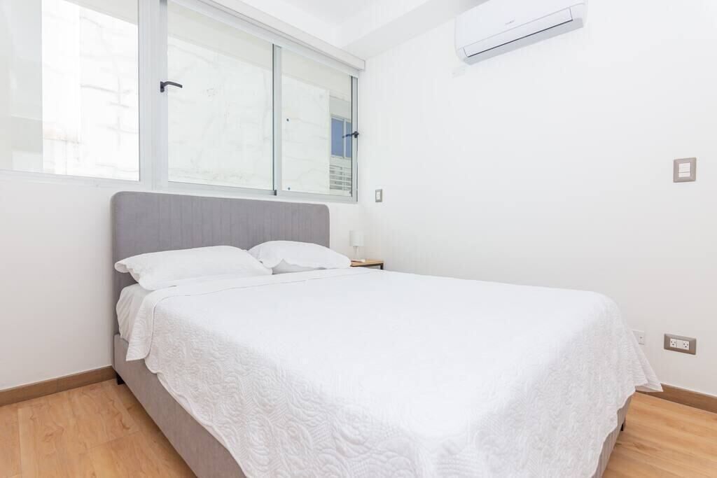 A neatly made bed in a room with a large window, a bedside table, and an air conditioning unit on the wall.