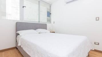 A neatly made bed in a room with a large window, a bedside table, and an air conditioning unit on the wall.