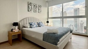 A modern bedroom with a large bed, a nightstand, a window with a city view, and abstract wall art.