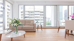 A modern living room with a beige sofa, a round white table, and a small side table with a vase of flowers. Large windows offer a view of high-rise buildings. - Luxury 1 bedroom apt free parking and balcony (Panamá)