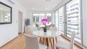 A modern living room with a round glass table, white chairs, a flat-screen TV, and a large window with a city view. - Luxury 1 bedroom apt free parking and balcony (Panamá)