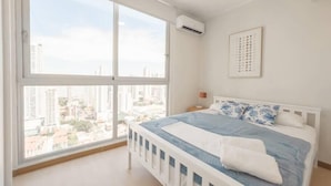 Ein Schlafzimmer mit großem Fenster, einem Bett mit blauer Bettwäsche, einem weißen Kopfteil und einem gerahmten Bild an der Wand.
