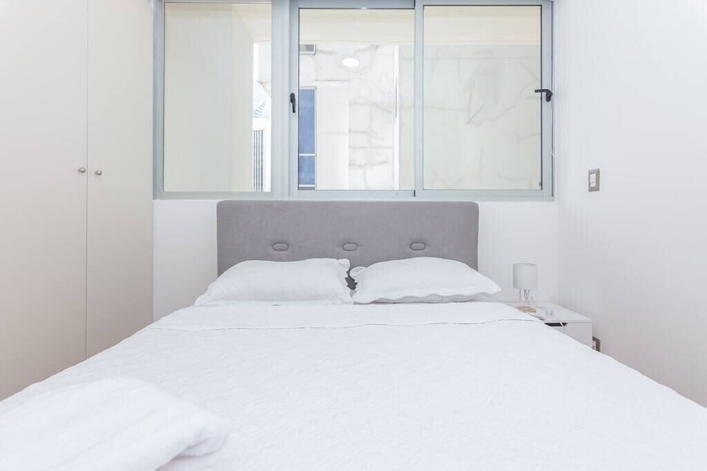 A neatly made bed with white linens, a grey headboard, and a bedside table with a lamp.