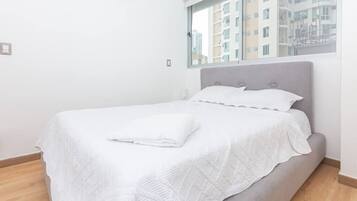 A neatly made bed with a white bedspread and pillows, a grey upholstered headboard, and a window with a view of buildings.
