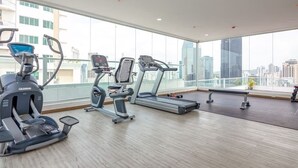 A modern gym with treadmills, elliptical machines, and a bench. Large windows offer a view of the city skyline.