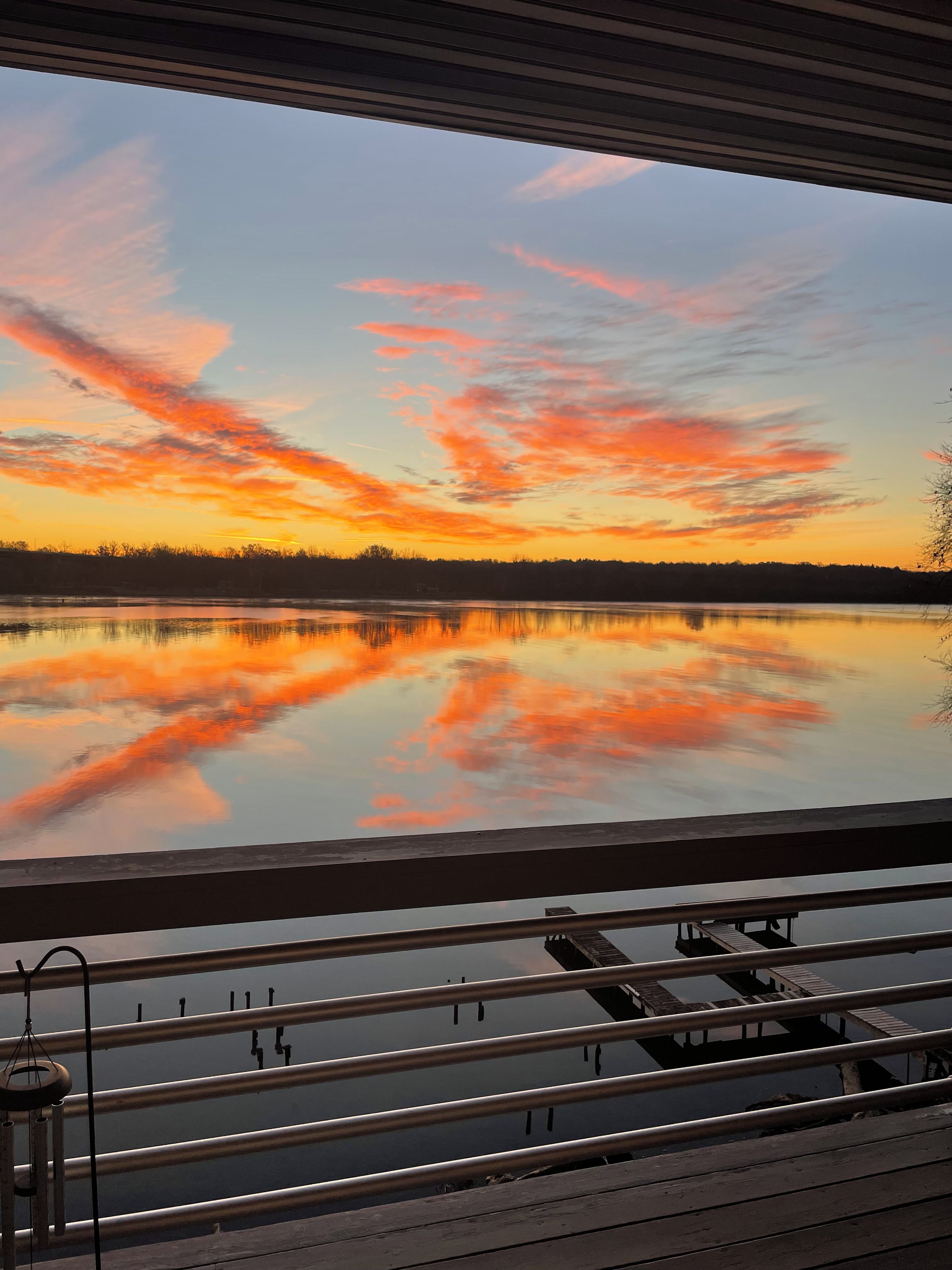 Näkymä ikkunasta, josta avautuu tyyni järvi, jonka yllä auringonlasku värjää taivaan, ja näkyvissä on myös laituri ja kaide.