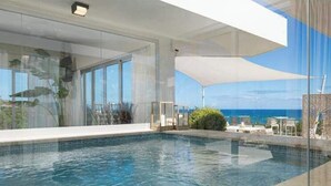 A modern indoor pool with clear glass walls, a view of the ocean, and a covered seating area. - Sunrise Suites (Puerto Plata)
