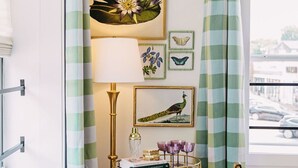 Une pièce avec un rideau à carreaux, une estampe botanique encadrée, une lampe, une petite table avec des livres et des lunettes, et une chaise.