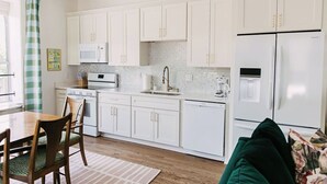 Une cuisine moderne avec des meubles blancs, un four à micro-ondes, une cuisinière et un réfrigérateur. On y trouve une table à manger avec des chaises, ainsi qu’un canapé vert dans l’espace salon.