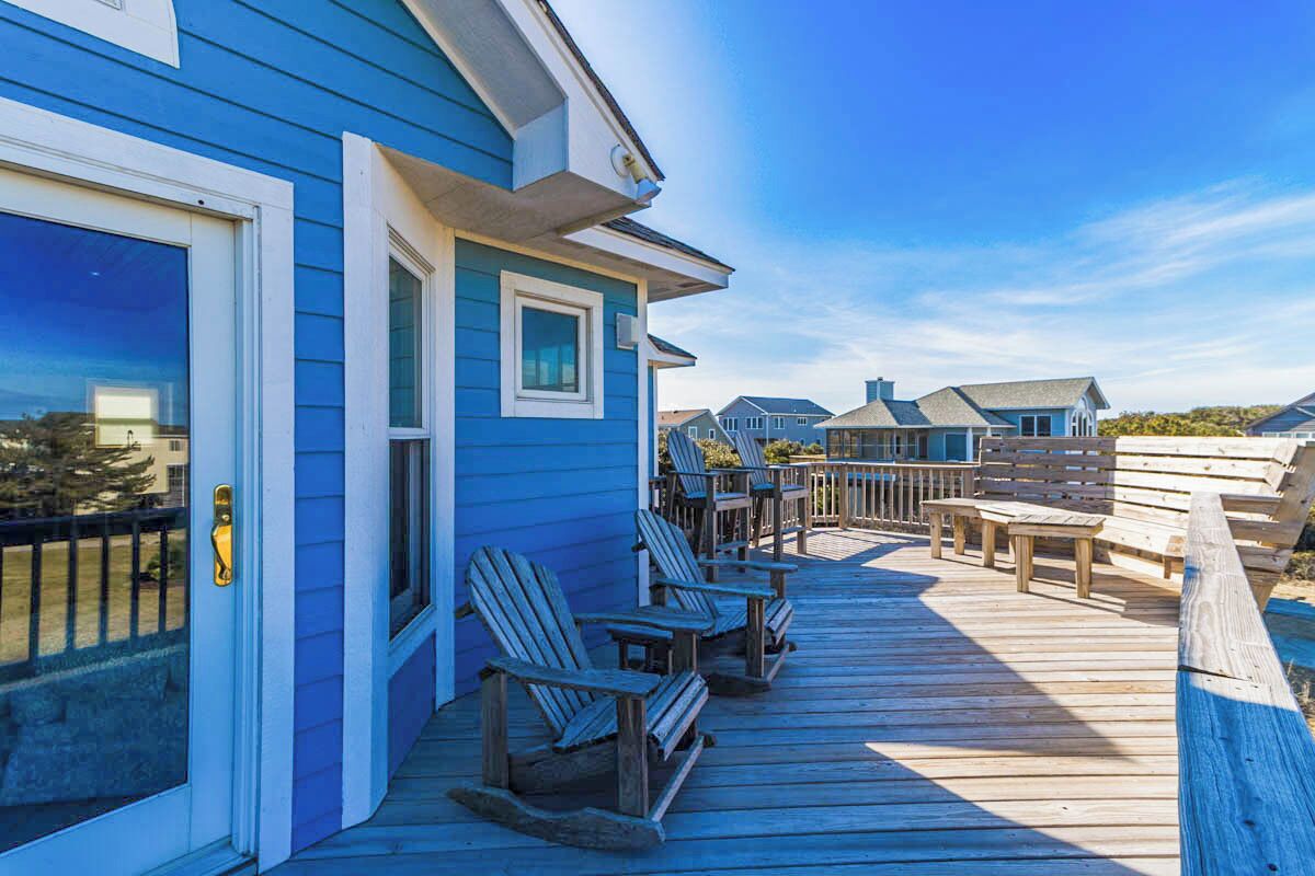 A wooden deck with Adirondack chairs and a table, leading to a blue house with a white door and windows.