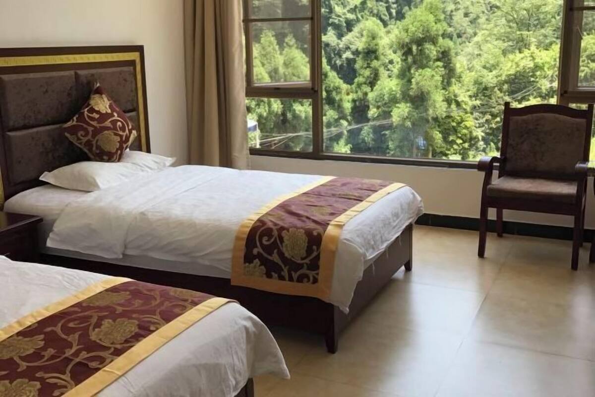 A hotel room with two beds, a large window with a forest view, and a chair.