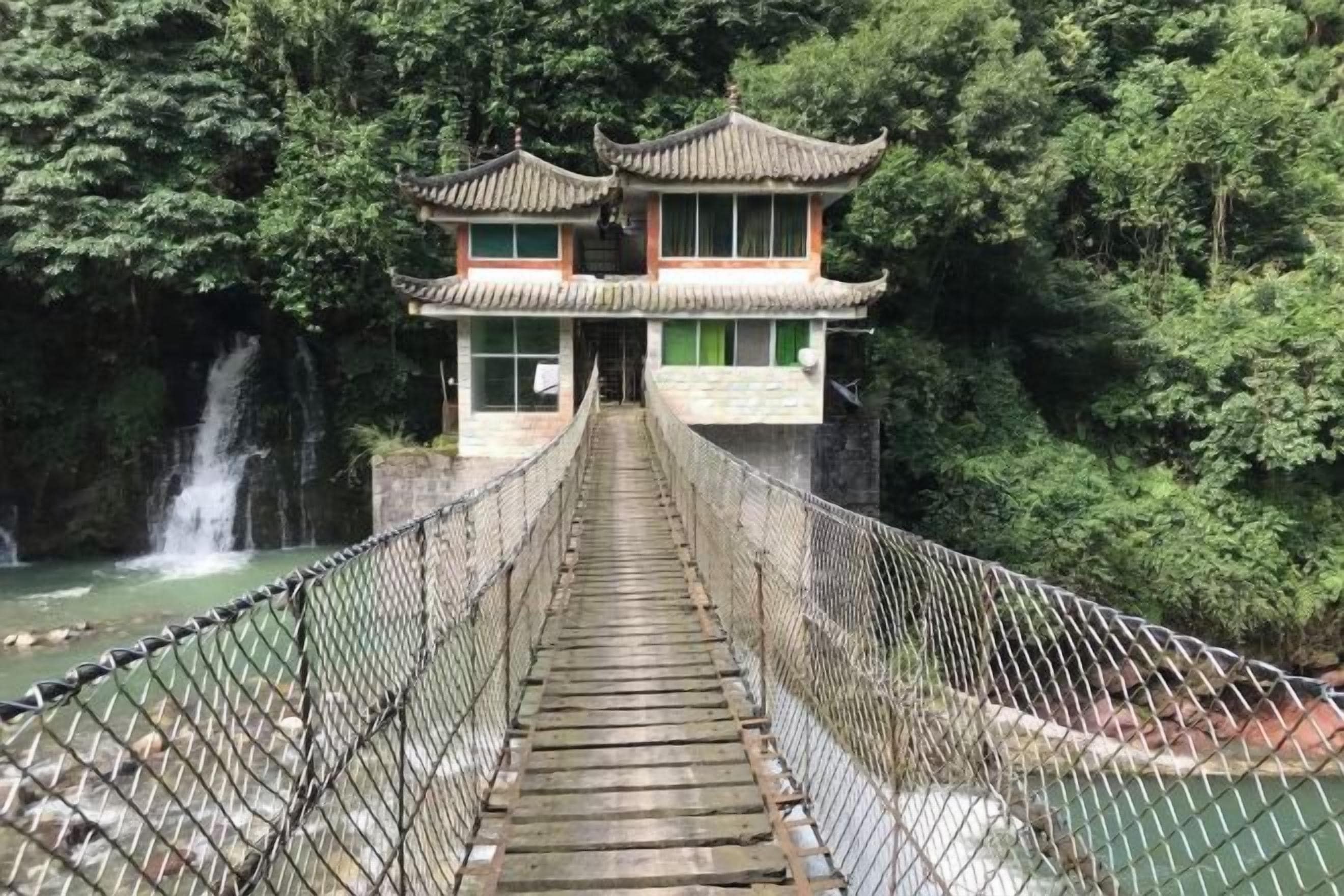Een hangbrug die leidt naar een traditioneel gebouw met een pannendak, omringd door weelderige begroeiing en een waterval.
